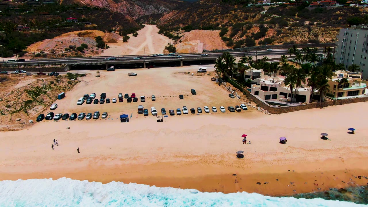Aerial frame at Punta Buzzards, Baja California Sur showing white surf at the bottom edge, scattered people and umbrellas on sand, rows of parked cars, a highway, palm trees, buildings, and dry mountains.