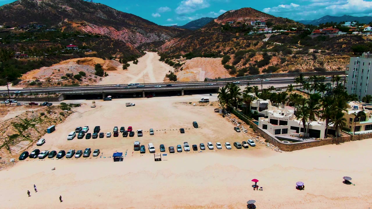 Drone view over sandy beach parking with highway and mountain slopes beyond.