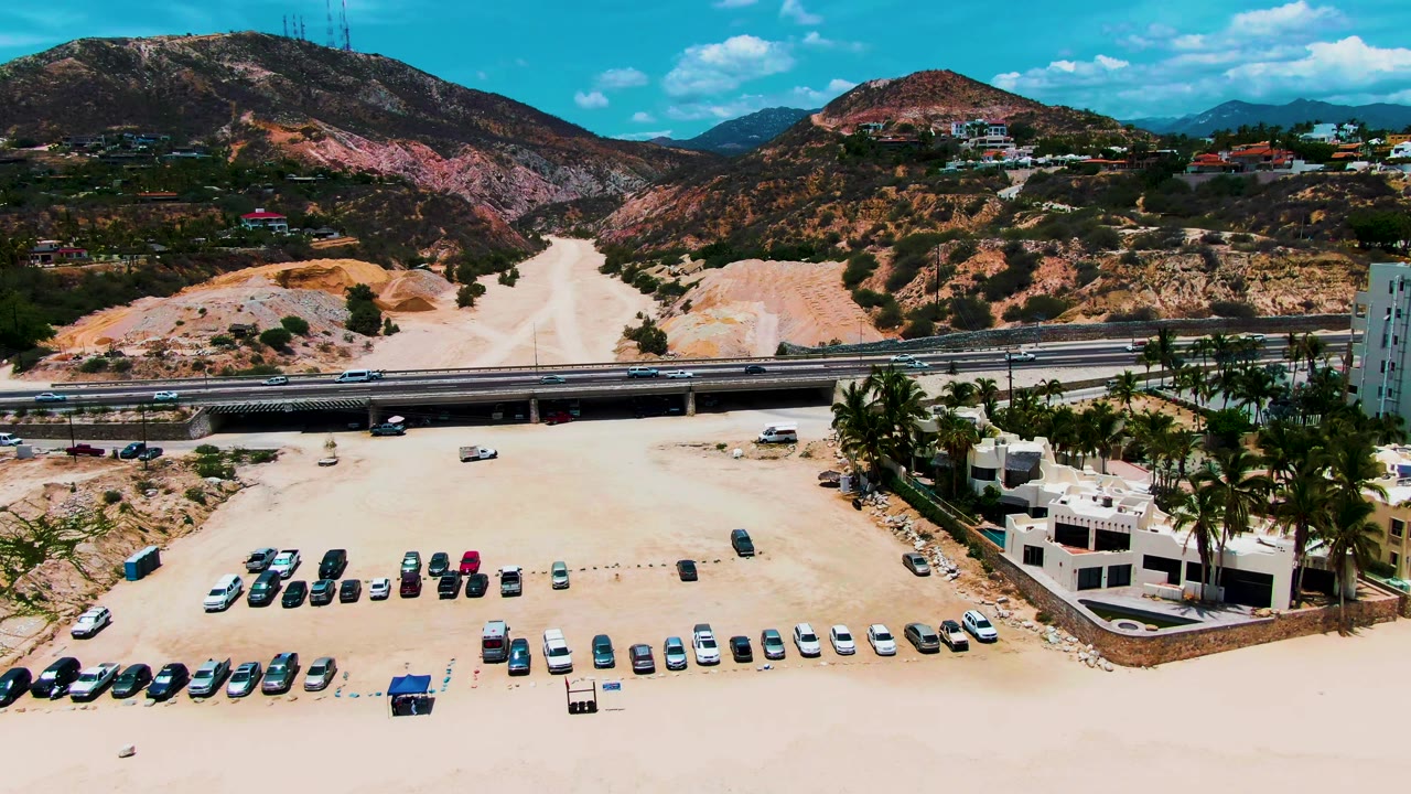 Punta Buzzards aerial frame showing an expanded view of the sandy parking area, rows of cars, small umbrellas near the lower beach, palm-lined buildings with a pool, highway traffic, and scrubby mountain ridges.