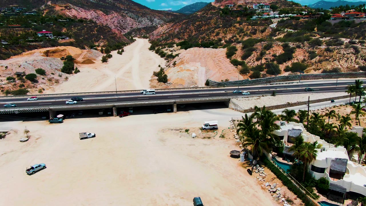 Aerial frame from Punta Buzzards, Baja California Sur showing a highway overpass over a dry sandy riverbed, hillside scrub, scattered homes, vehicles, palms at right, and no visible people.