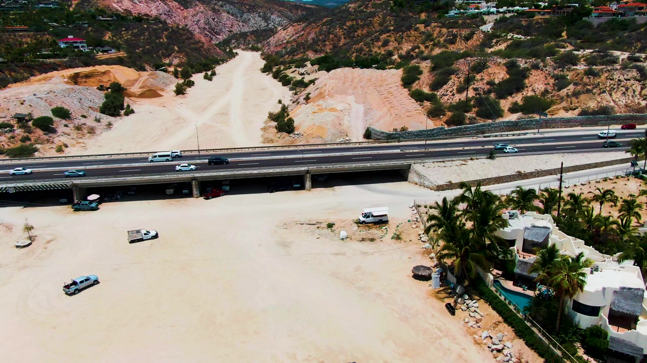 Aerial view in Punta Buzzards, Baja California Sur with cars on the highway overpass, a broad pale riverbed below, white residential buildings and palms at right, and no visible people.