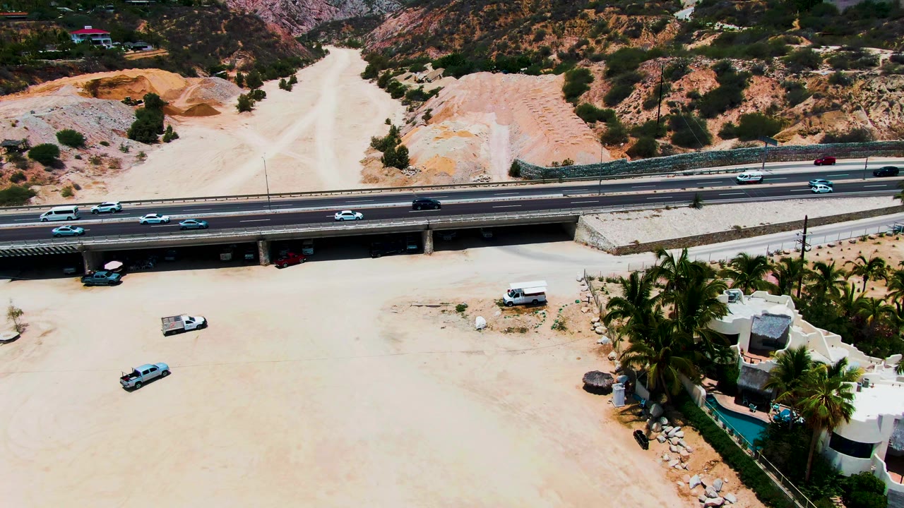 Punta Buzzards aerial frame showing highway vehicles crossing the overpass, a dry sandy wash beneath, hillside tracks in the background, residential buildings at right, and no visible people.