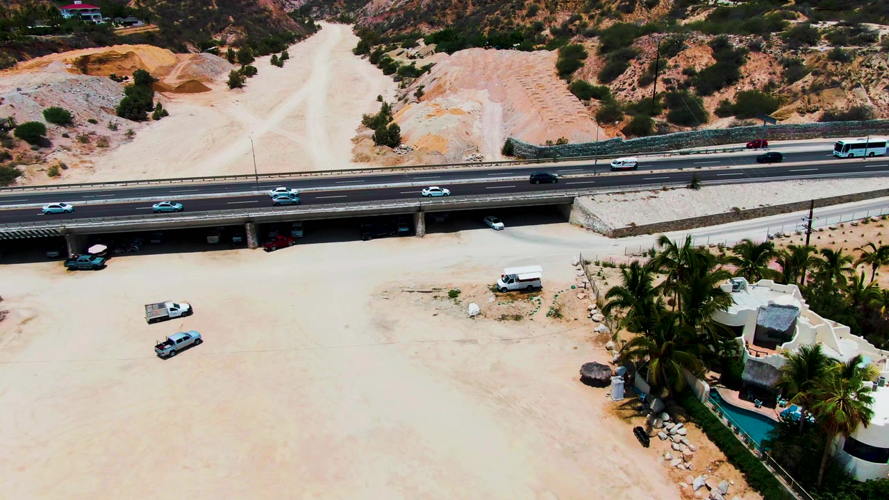 Aerial shot of highway traffic over a dry riverbed with homes and palms at the lower right.