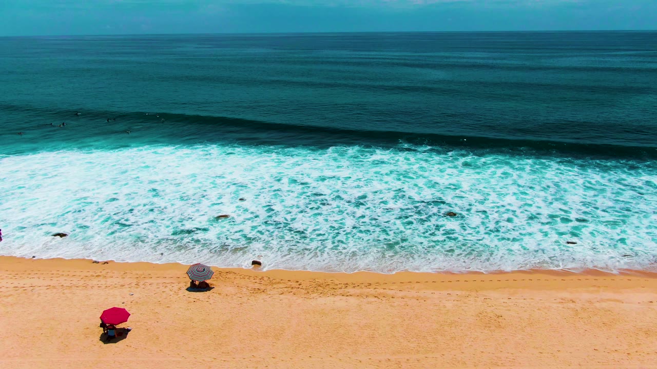 Aerial view of Buzzards Beach in Punta Buzzards, Baja California Sur, with white surf reaching golden sand and two separated umbrellas, no visible people.