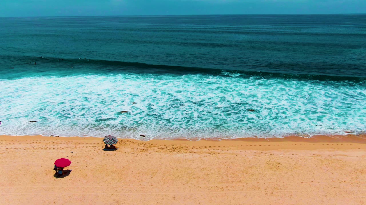 Buzzards Beach shoreline from above, with a broad white foam line closer to the sand and the red and gray umbrellas still fixed on the beach, no visible people.