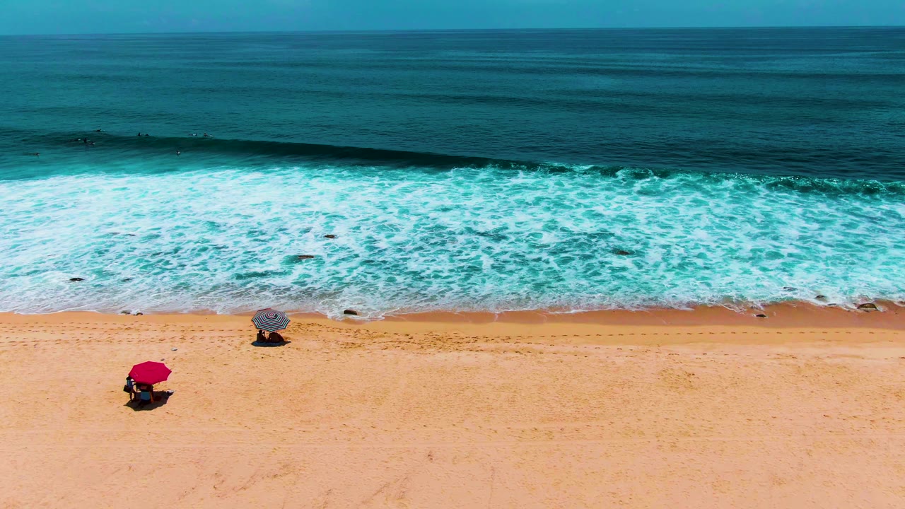 High aerial frame over Punta Buzzards beach, showing darker blue water beyond a bright foam band and two umbrellas spaced along the sandy shore, no visible people.