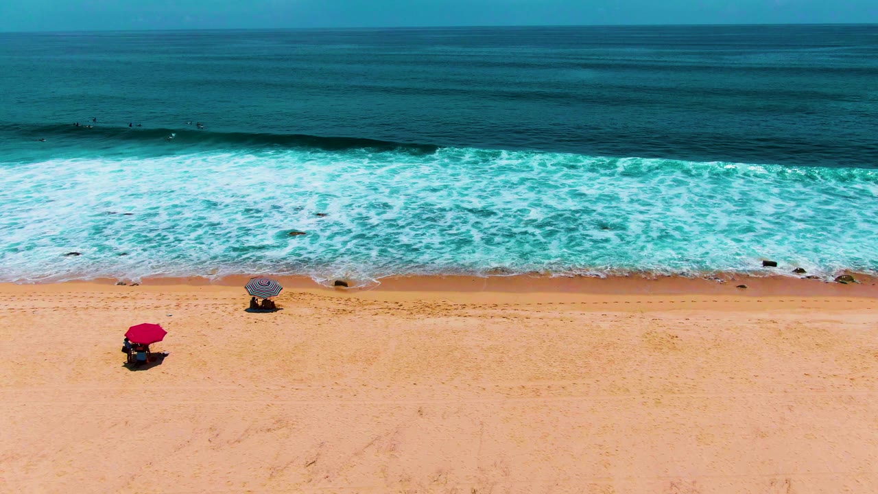 Aerial beach scene with surf washing toward umbrellas on sand.