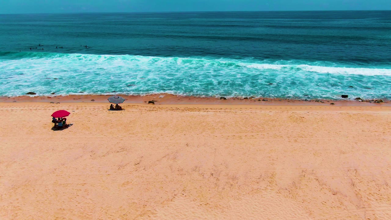 Wide view of Buzzards Beach in Punta Buzzards, Baja California Sur, with pale sand, a red umbrella at left, a gray umbrella near center, small visible people, white surf, and a blue ocean horizon.