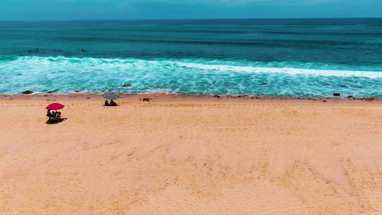 Beach umbrellas and small people below breaking waves and a level ocean horizon.