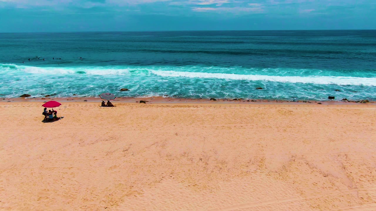 A high beach view at Punta Buzzards shows a red umbrella near the lower right, small visible people on the sand, a gray umbrella farther left, white surf, and layered blue ocean water.