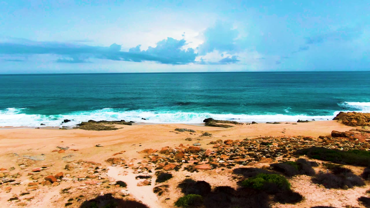 Aerial view of sandy beach, rocks, turquoise surf, and cloudy ocean horizon.