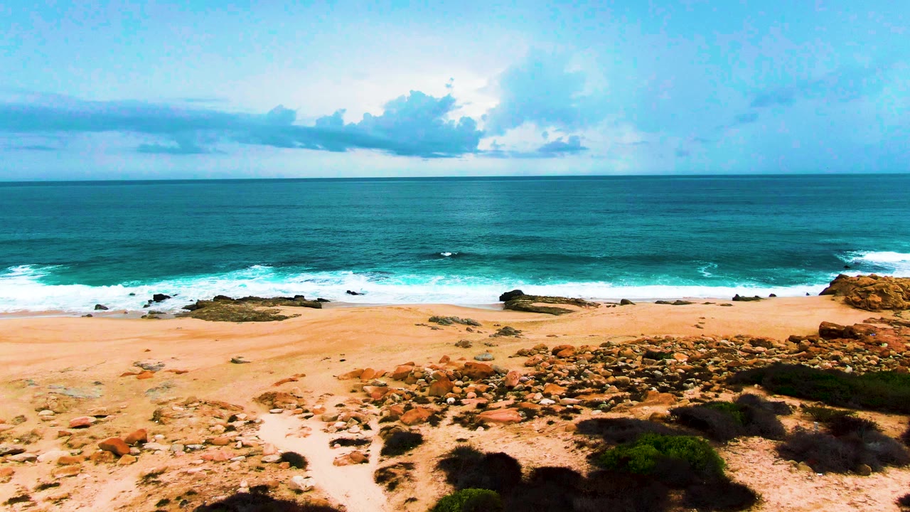 Aerial frame from Punta Buzzards showing tan sand across the lower frame, dark rock patches near the waterline, blue-green ocean, white breaking waves, and no visible people.