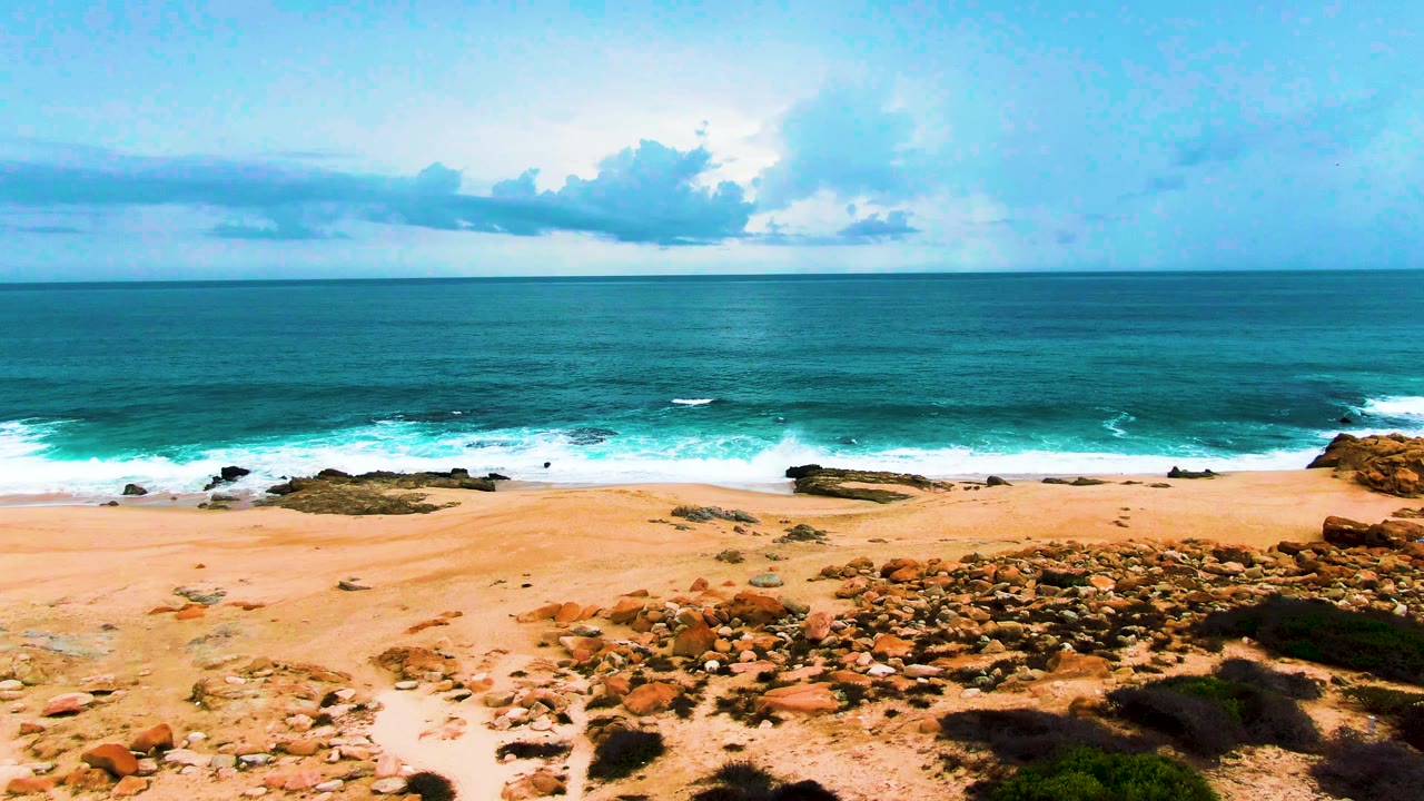Punta Buzzards shoreline with a broad sandy foreground, a cluster of orange-brown rocks to the right, turquoise water rolling toward shore, cloud cover above the horizon, and no visible people.