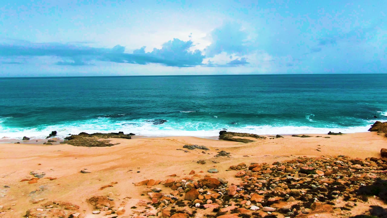 High aerial view over Punta Buzzards with pale sand and low rocks in the foreground, white surf curling across the shoreline, a flat ocean horizon, and no visible people.