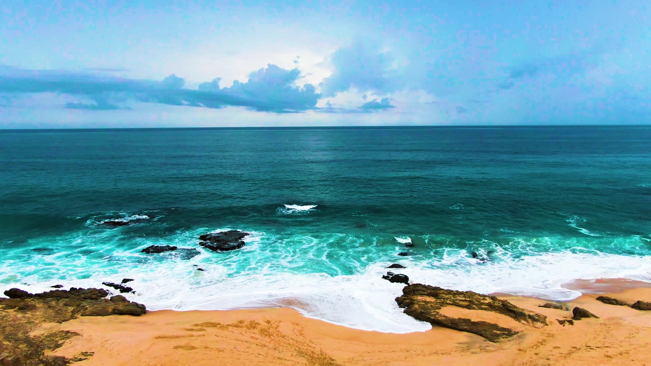 Aerial frame from Punta Buzzards, Baja California Sur, showing white surf wrapping around dark rocks beside tan sand, with open blue ocean and no visible people.