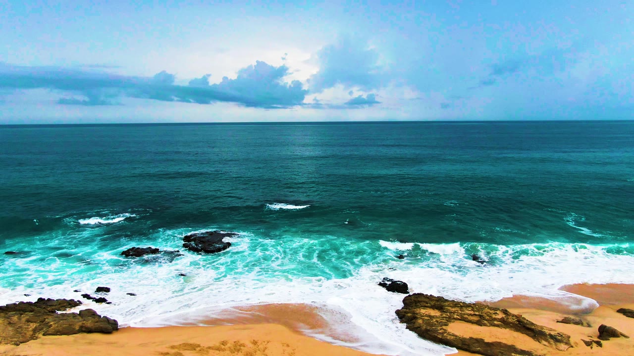 Aerial view at Punta Buzzards with more whitewater spread across the lower shoreline, dark rock outcrops in the surf, a level horizon, and no visible people.