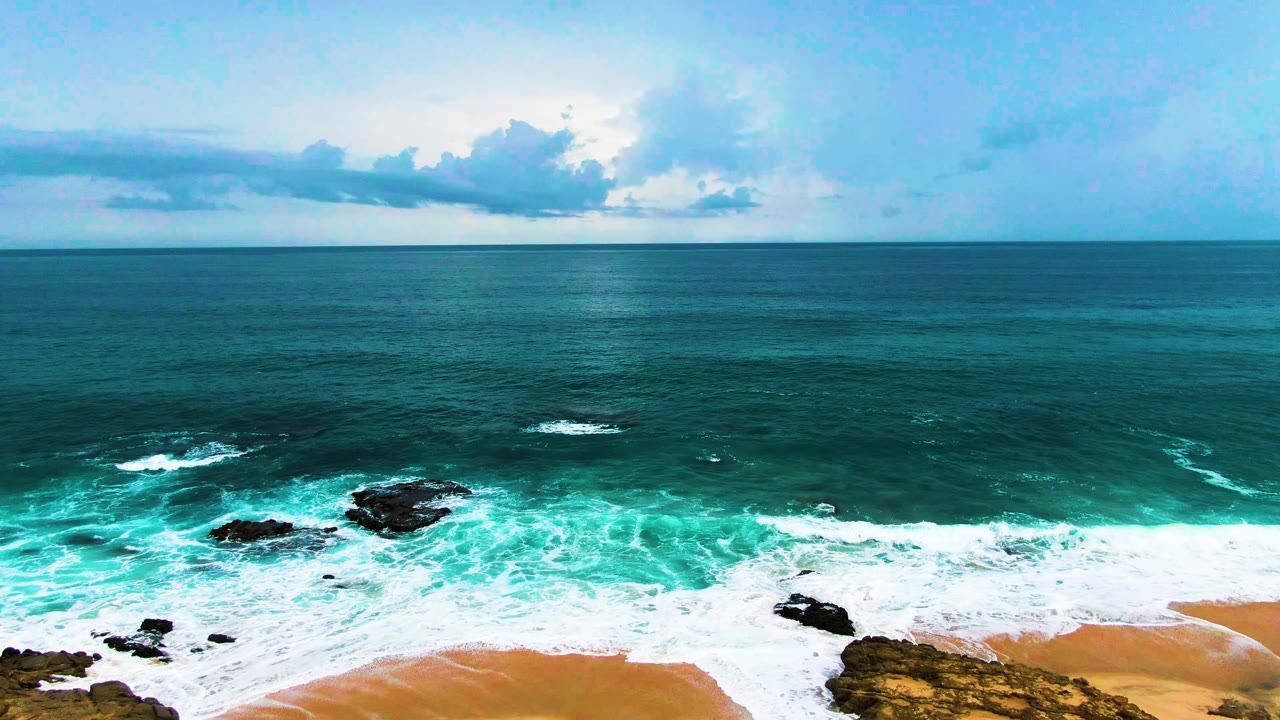 Punta Buzzards shoreline frame with a broad band of turquoise surf, scattered black rocks, a small sandy curve at the bottom, and no visible people.