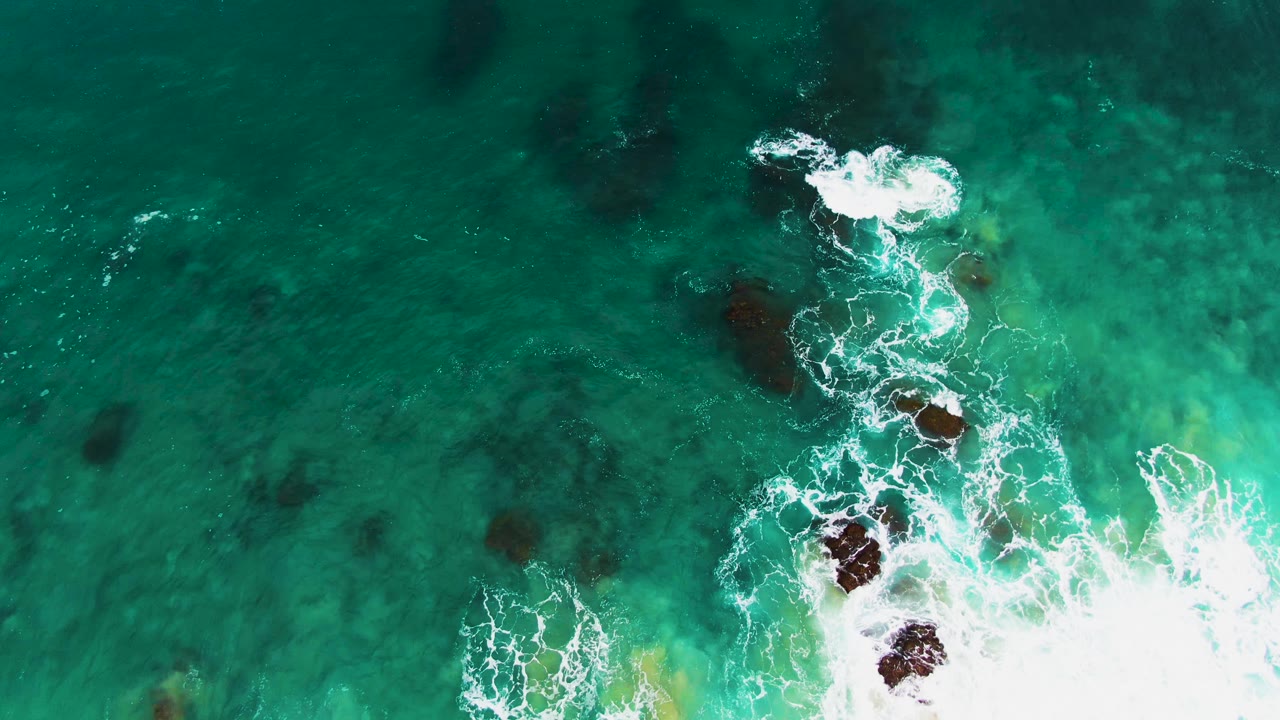 Overhead view at Buzzards Beach East Cape, Baja California Sur, showing turquoise water and white surf breaking around dark submerged rocks near the lower frame, with no visible people.