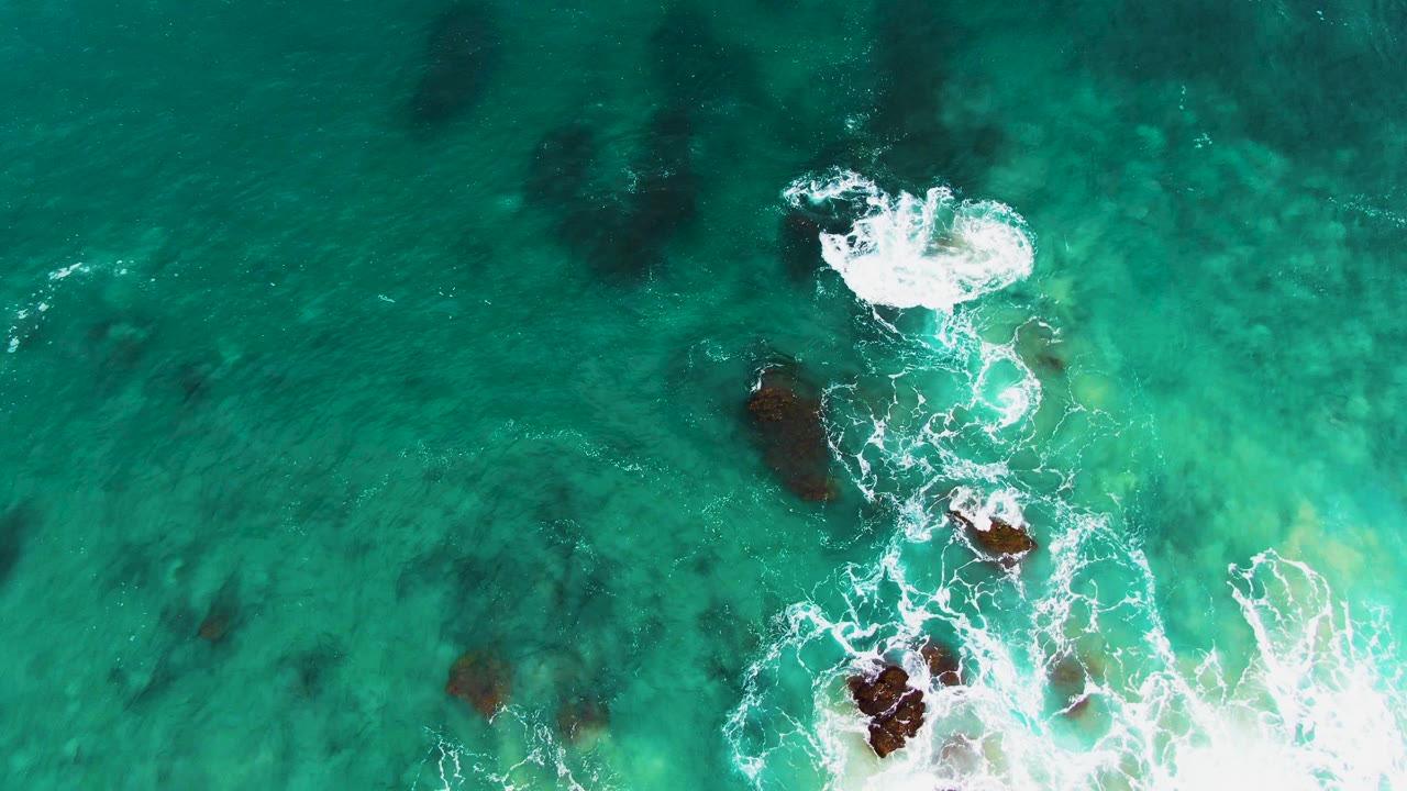 A top-down Buzzards Beach East Cape frame shows a larger white foam burst over the lower rock cluster, surrounded by clear turquoise water and darker underwater patches, with no visible people.