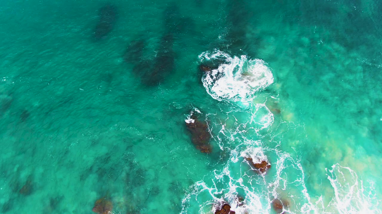 The third overhead frame at Buzzards Beach East Cape shows foam trails extending down from a top rock break toward darker rocks below, all within clear turquoise water and no visible people.