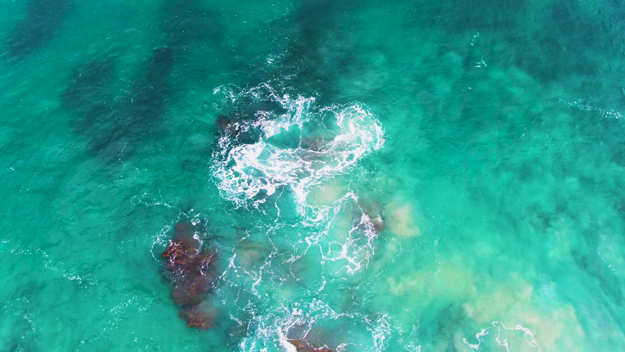 Overhead view at Punta Buzzards, Baja California Sur, showing turquoise water, dark submerged rocks, white foam beginning to curl around the upper rock cluster, and no visible people.