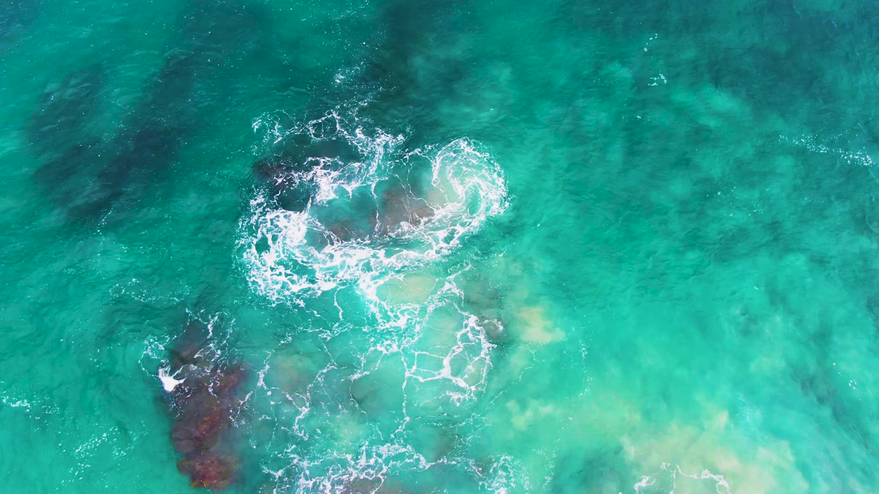 Overhead frame at Punta Buzzards, Baja California Sur, with white foam forming a loose ring over submerged rocks while darker rock shapes show through clear turquoise water and no visible people.