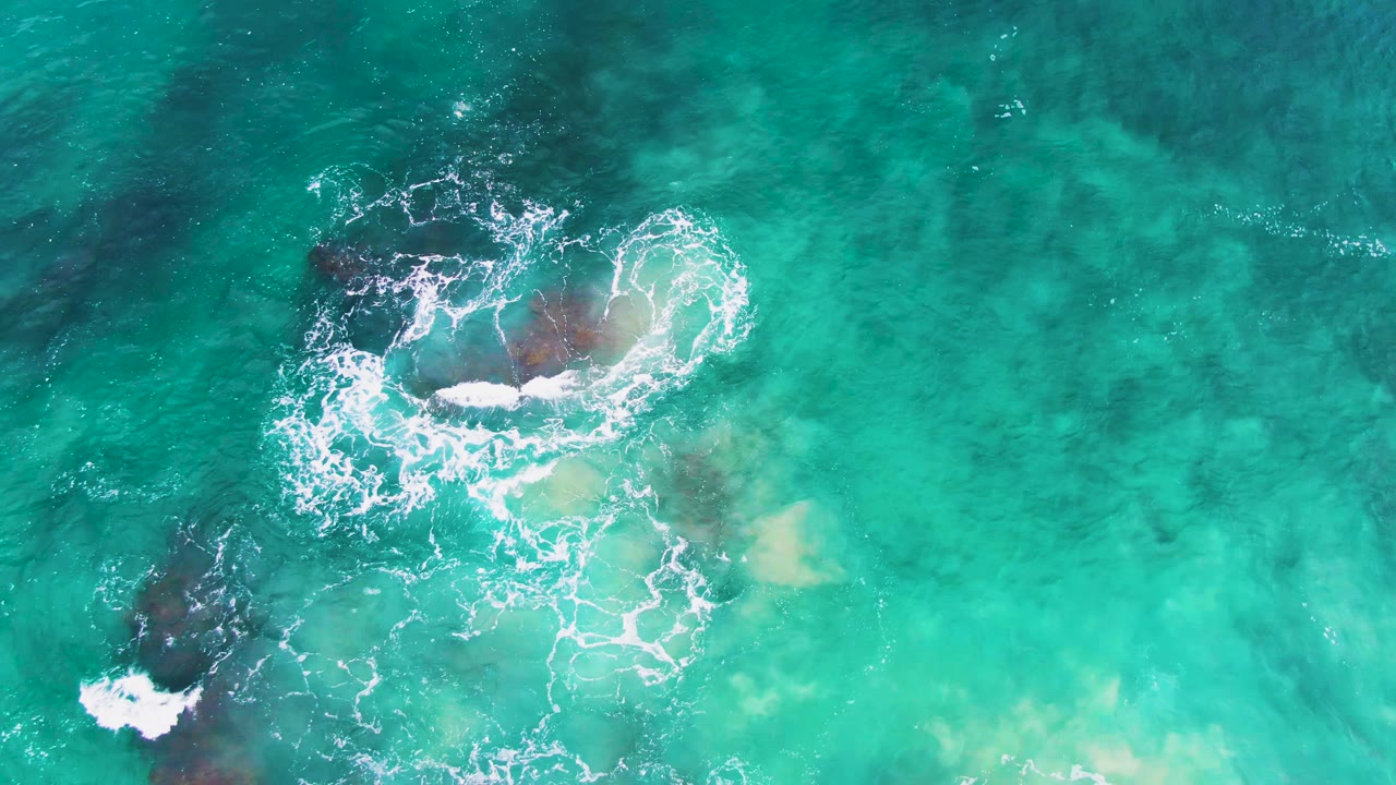 Overhead view at Punta Buzzards, Baja California Sur, where a brighter band of foam crosses the upper rock cluster, a darker rock sits lower left, and no visible people appear in the water.