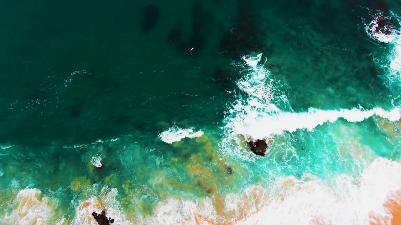 Top-down aerial view at Punta Buzzards as white surf pushes diagonally through blue-green water around exposed rocks, with no visible people.