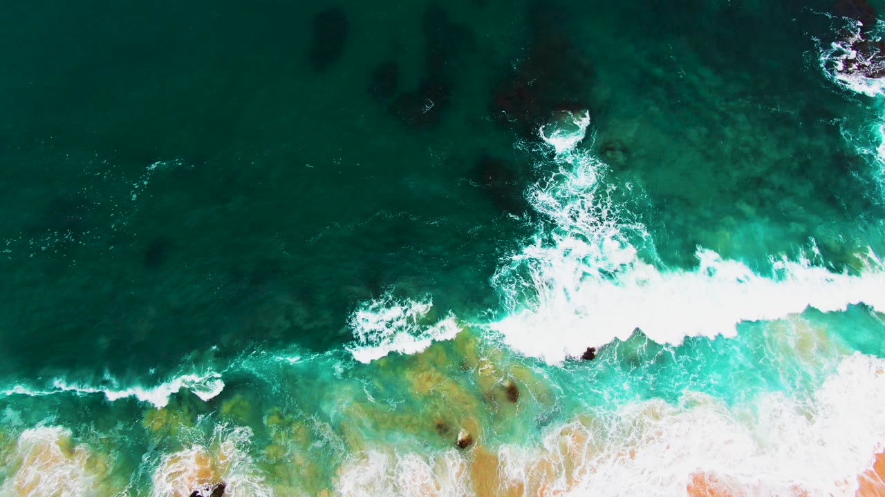 Aerial view at Punta Buzzards showing a thicker white breaker crossing the lower half of turquoise shallows, dark rocks, and no visible people.
