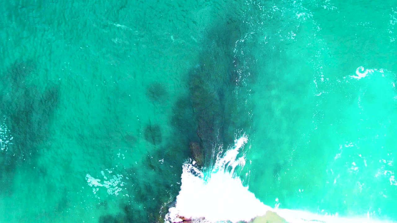 Aerial frame from Punta Buzzards with a darker rock band beneath turquoise water, white surf foam along the lower left, and no visible people.