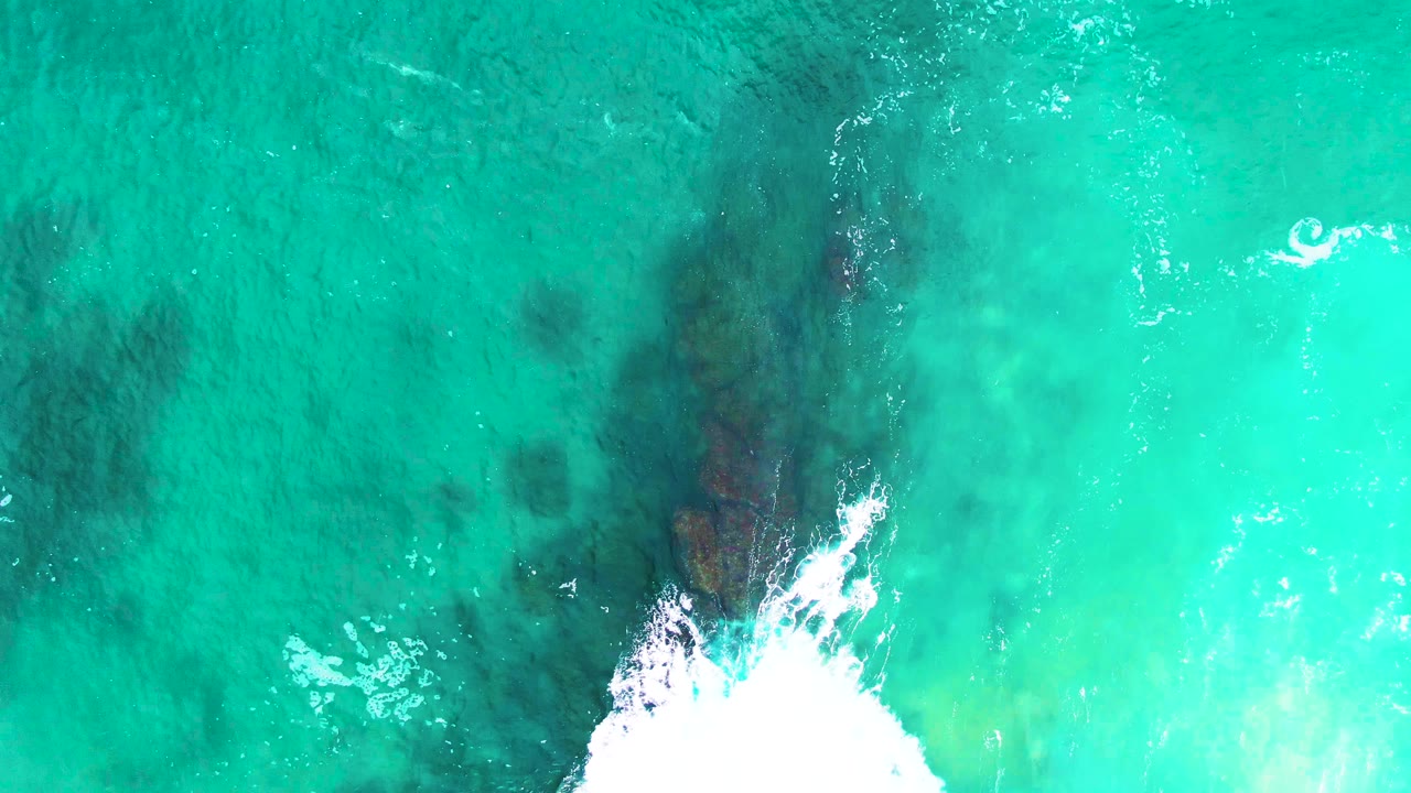 Overhead Punta Buzzards water view with a broad dark rock shape below the surface, foamy surf near the bottom, small bright glints, and no visible people.