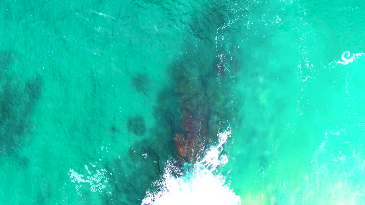 Aerial view at Punta Buzzards showing the rock formation more centered under clear turquoise water, white foam at the lower edge, and no visible people.