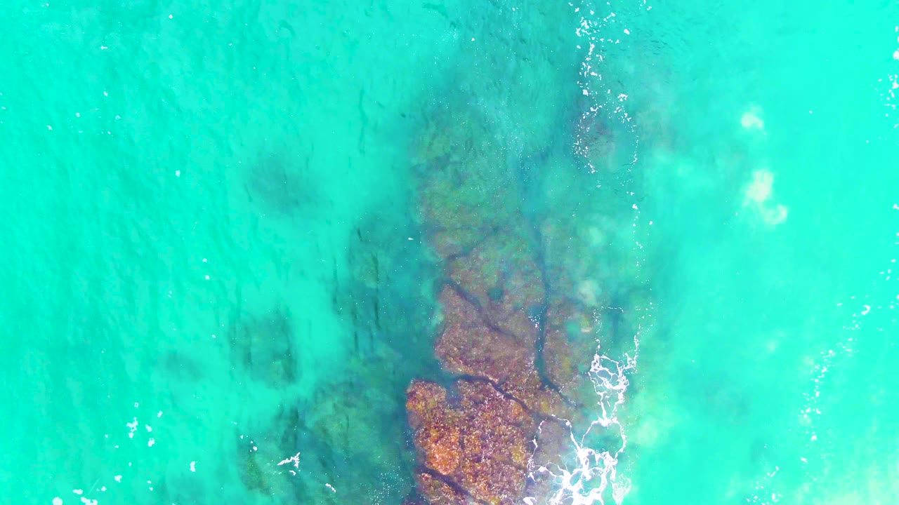 Top-down view at Punta Buzzards with reddish-brown rocks visible beneath turquoise water, light foam near the rock edge, and no visible people.