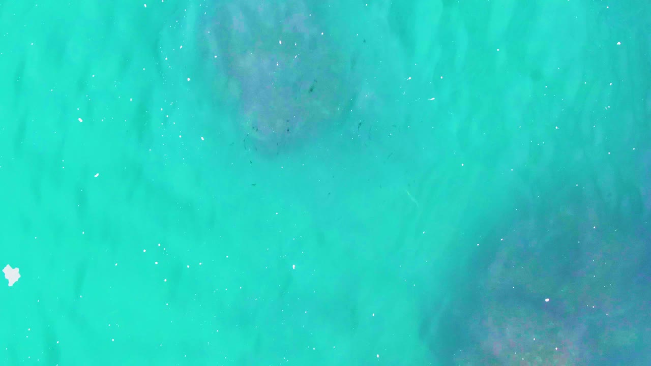 Turquoise Punta Buzzards water shows a darker submerged patch near the middle, faint ripple lines, drifting white specks, and no visible people.