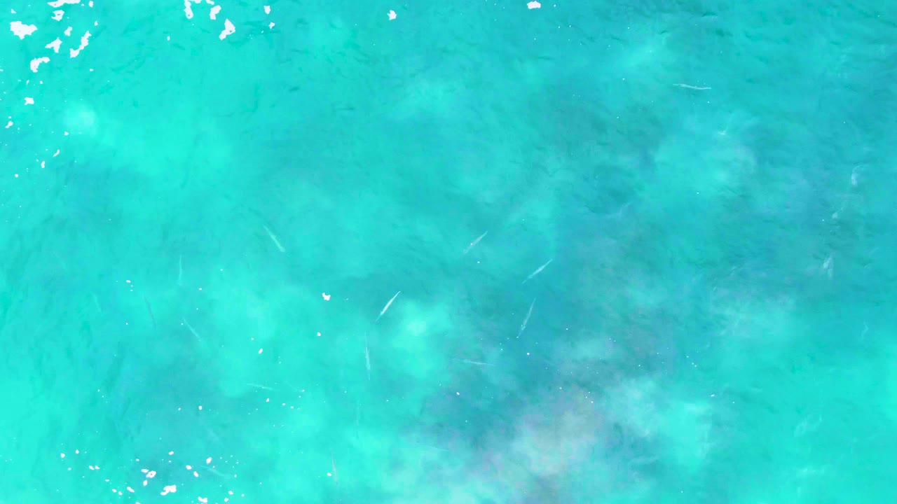 Clear turquoise ocean surface at Punta Buzzards shows darker seabed areas beneath the water, scattered bubbles near the top edge, and no visible people.