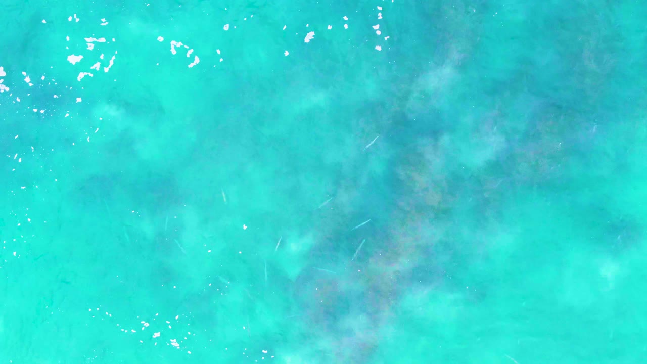 Straight-down view of turquoise shallow water at Buzzards Beach in Baja California Sur, with scattered white foam flecks, muted darker underwater patches, and no visible people.