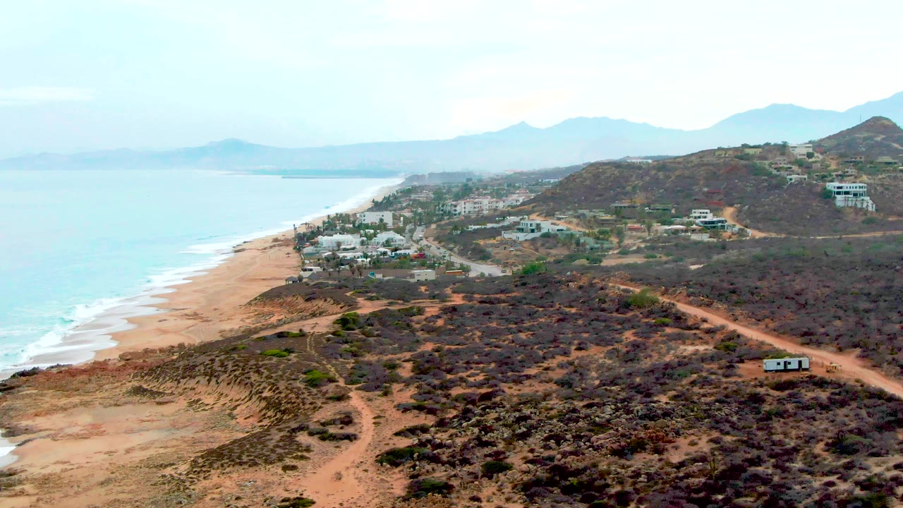 Wide coastal aerial with beach on the left and arid hillside roads on the right.