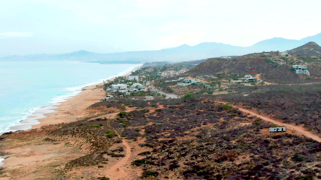 Aerial view over Buzzards Beach with turquoise water at left, sandy shoreline, dark scrub-covered hills, dirt roads, small buildings, hazy mountain ridges, and no visible people.