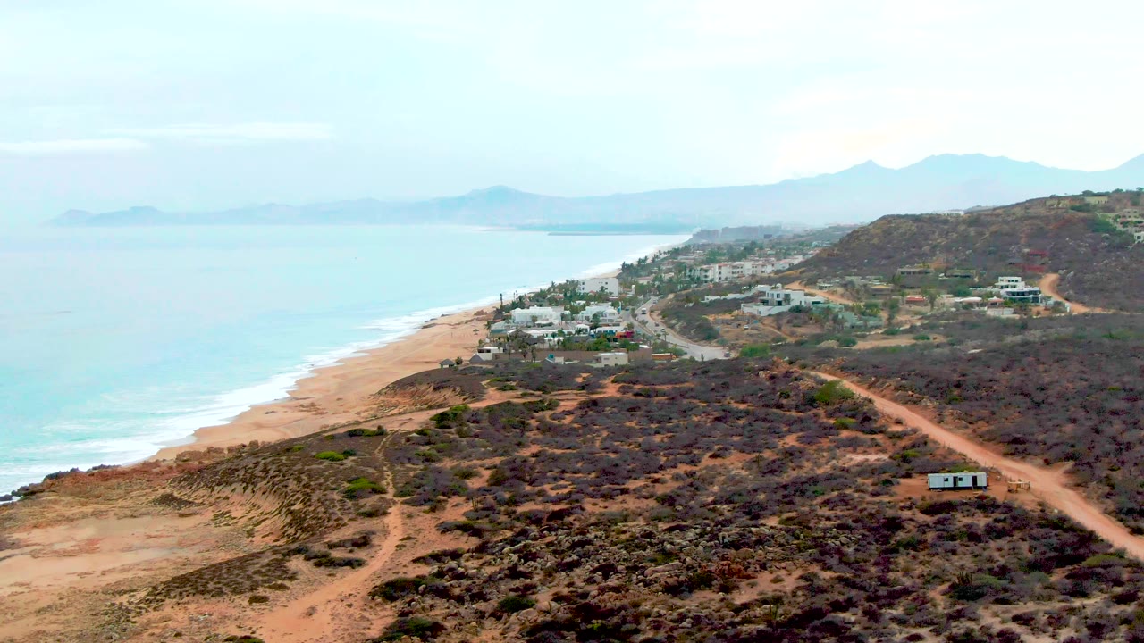 At 0:00, an aerial view over Punta Buzzards shows the beach and blue ocean to the left, dry scrub hills and a dirt road to the right, distant mountains on the horizon, and no visible people.