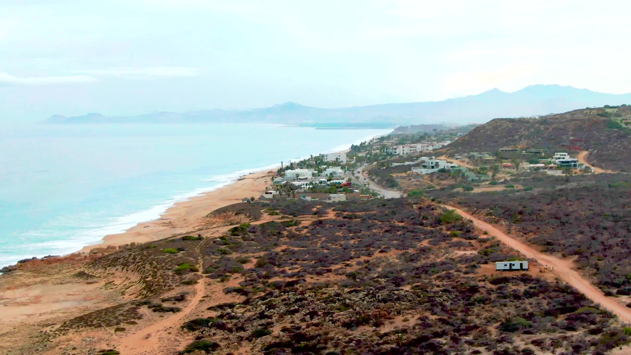 At 0:01, the Punta Buzzards coastline remains in an elevated view, with a winding dirt road crossing dry brush toward small white buildings near the beach and no visible people.