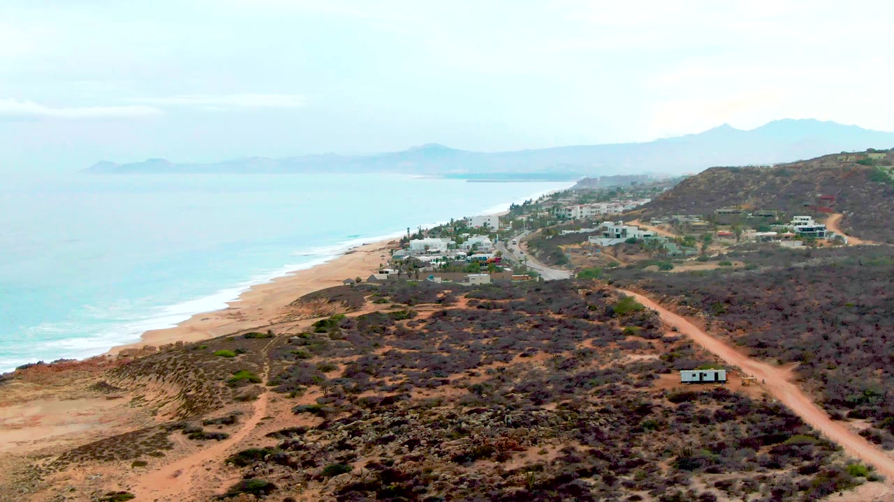 At 0:02, the aerial composition shows turquoise water and a sandy beach on the left, dirt roads cutting through Punta Buzzards scrub hills, scattered houses, and no visible people.