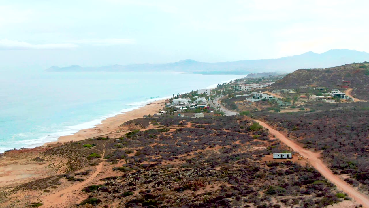 Aerial coastal landscape with beach, pale blue sea, dry hillside roads, and scattered buildings.