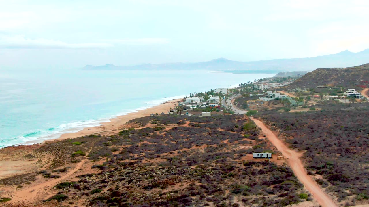 At 0:00, an elevated view over Punta Buzzards shows a dirt road through dry scrub near a sandy beach, turquoise ocean, scattered coastal buildings, distant hills, and no visible people.