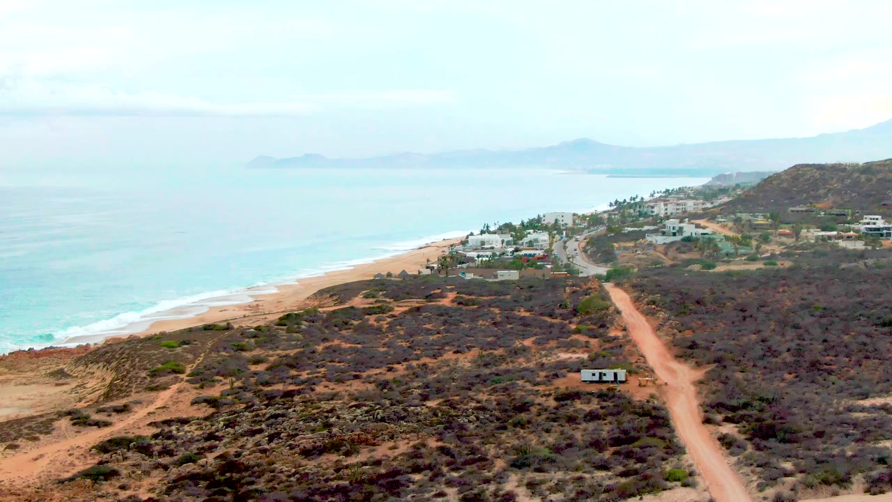 At 0:02, the road appears slightly farther right in the frame, with dark scrub foreground, beachside structures, blue-green water, distant landforms, and no visible people.