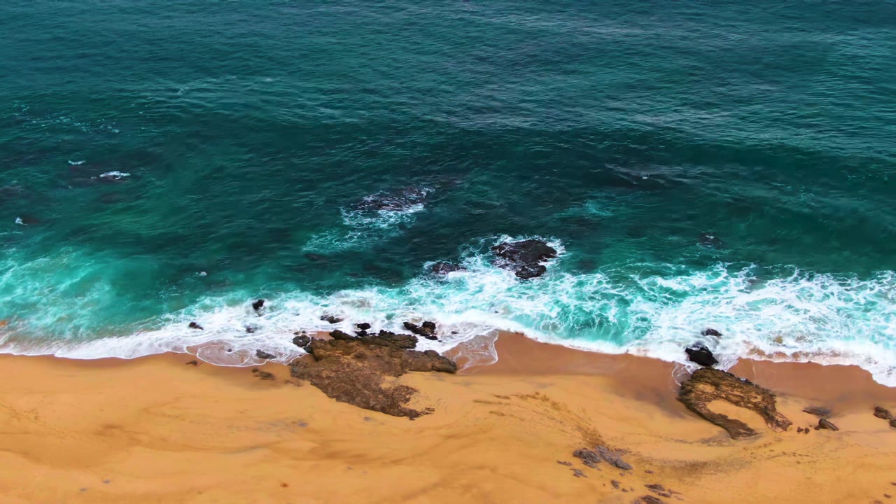 At Punta Buzzards in Baja California Sur, turquoise water pushes white foam toward dark shoreline rocks and golden sand, with no visible people.