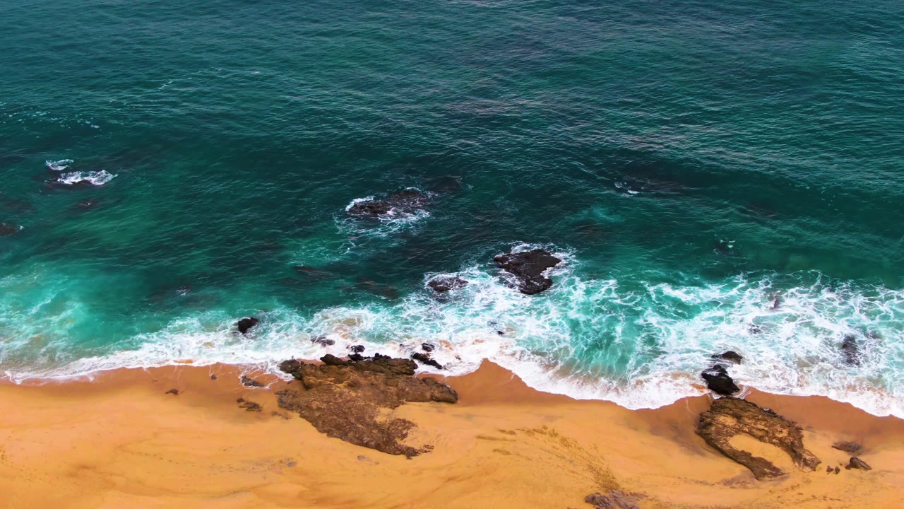 At Punta Buzzards in Baja California Sur, a fresh band of foam wraps around dark rocks while the sandy beach stays visible below the break, with no visible people.
