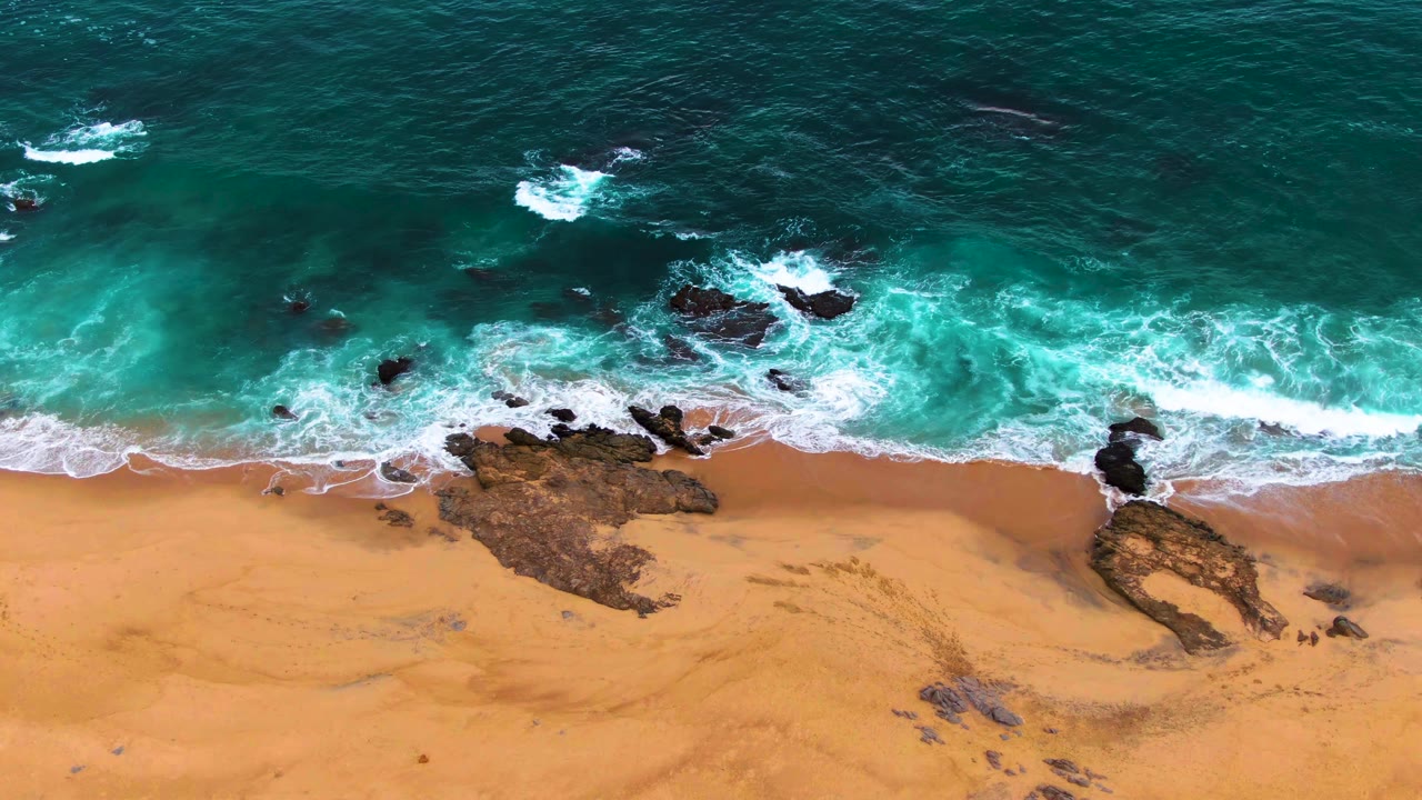 Aerial frame from Punta Buzzards, Baja California Sur, showing turquoise waves meeting a tan sandy beach with dark rocks near the surf line and no visible people.