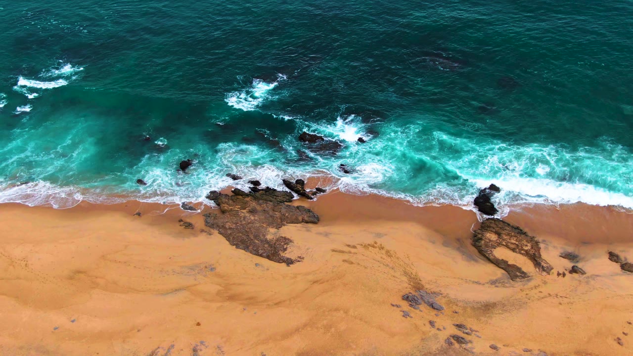 Aerial frame from Punta Buzzards, Baja California Sur, with whitewater wrapping around several dark rocks as blue-green water reaches the sandy shore and no visible people.