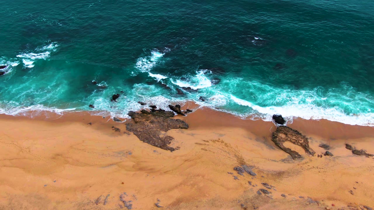 Aerial frame from Punta Buzzards, Baja California Sur, showing a brighter band of surf crossing dark rocks between deep teal water and tan sand with no visible people.
