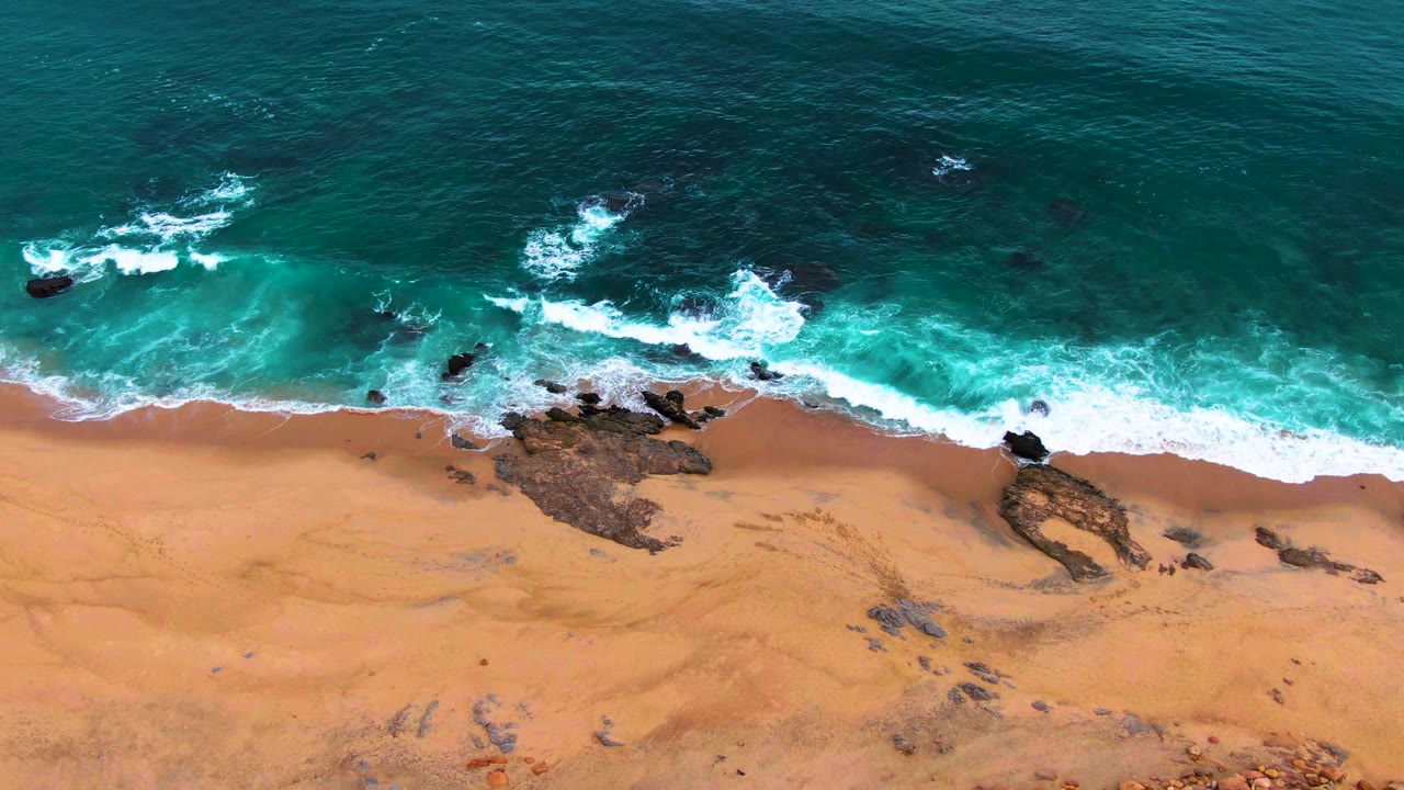 Wide aerial beach view with waves, rocks, and open sand.